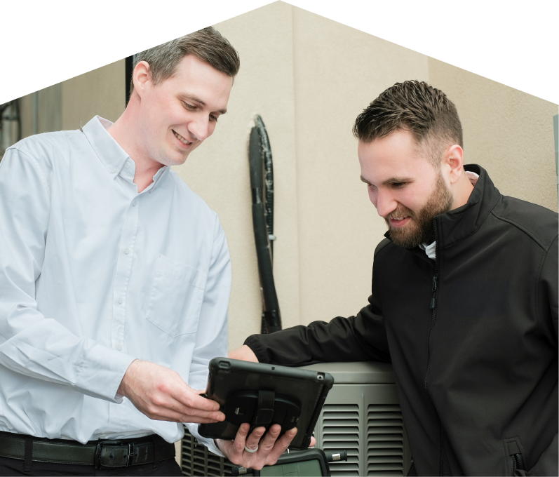 A Furnasman technician in a white shirt shows service details on a tablet to a smiling client in a black jacket, standing beside an AC unit outside a beige building. A Furnasman technician in a white shirt shows service details on a tablet to a smiling client in a black jacket, standing beside an AC unit outside a beige building.
