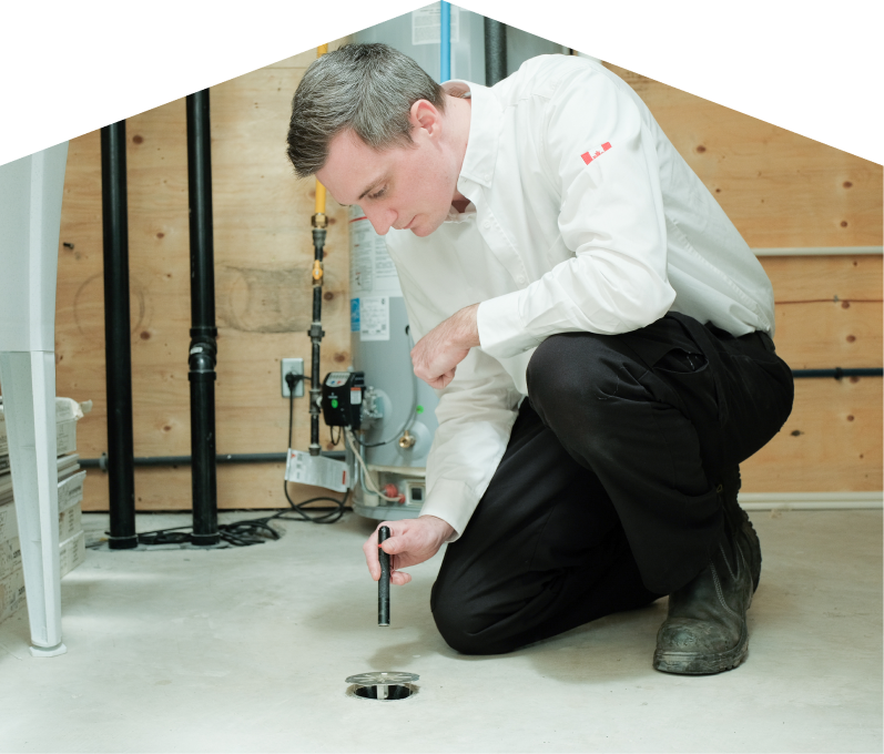 Furnasman plumbing specialist inspecting a basement floor drain with a flashlight during a drain cleaning service, in a utility room with visible pipes, water heater, and wood panel walls. Furnasman plumbing specialist inspecting a basement floor drain with a flashlight during a drain cleaning service, in a utility room with visible pipes, water heater, and wood panel walls.