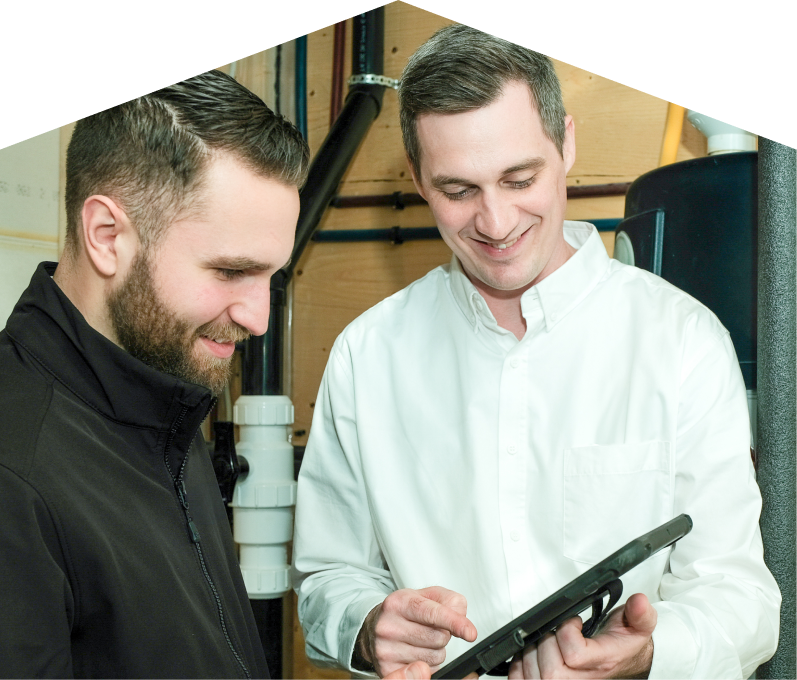 Furnasman specialist reviewing maintenance plan options with a smiling homeowner using a tablet, inside a utility room with visible piping. Furnasman specialist reviewing maintenance plan options with a smiling homeowner using a tablet, inside a utility room with visible piping.