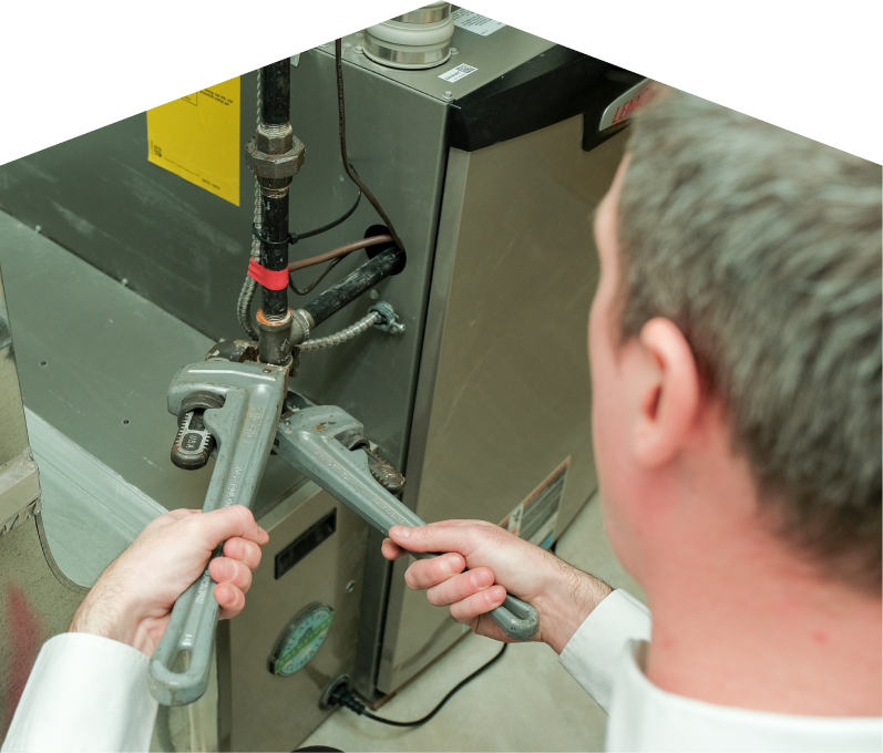 Furnasman plumbing specialist using two pipe wrenches to service a gas line connection beside a residential heating unit.