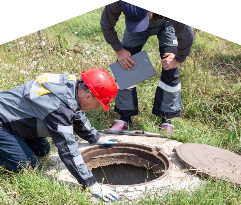 Furnasman sewer crew inspecting an open manhole in a grassy field, with one member documenting findings on a clipboard. Furnasman sewer crew inspecting an open manhole in a grassy field, with one member documenting findings on a clipboard.