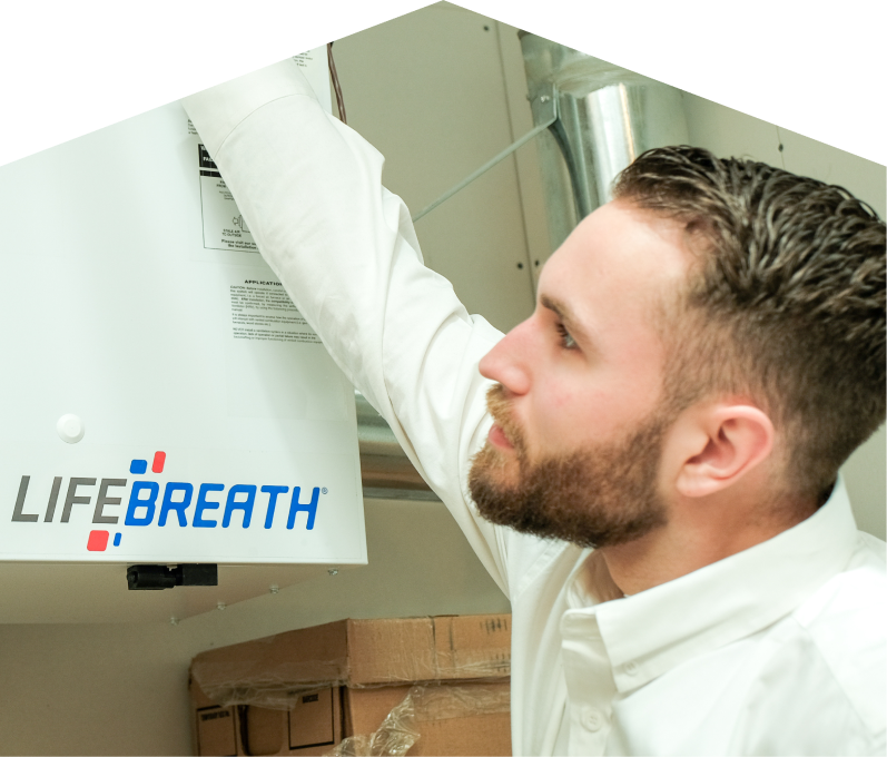 Furnasman indoor air quality specialist inspects a Lifebreath ventilation unit related to UV air purifiers, with ductwork and boxes in the background.