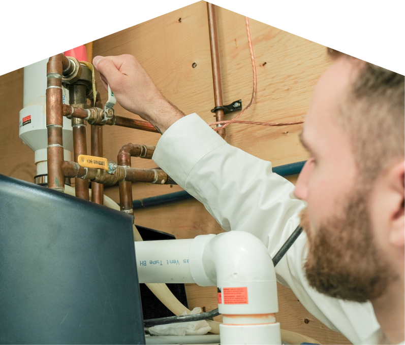 Furnasman water heater specialist tightens a valve on copper piping during a water heater installation, with exposed wood paneling and wiring in the background. Furnasman water heater specialist tightens a valve on copper piping during a water heater installation, with exposed wood paneling and wiring in the background.